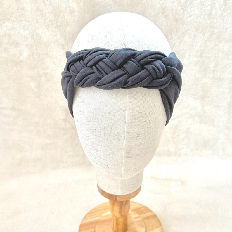 May include: A dark gray fabric headband featuring a thick, braided design that wraps around the crown. The headband is displayed on a plain, off-white mannequin head, showcasing the uniform color and the intricate, woven texture of the material.