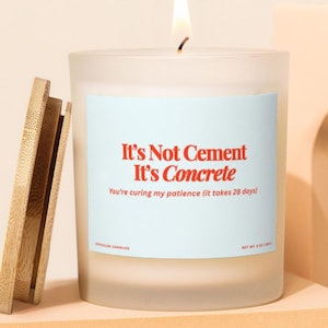以下が含まれることがあります： 点灯した、木製の蓋が付いた、つや消しガラスのキャンドル。キャンドルには、赤色の「It's Not Cement It's Concrete」というテキストが書かれた水色のラベルが付いています。下のテキストは「You're curing my patience (it takes 28 days)」と書かれています。