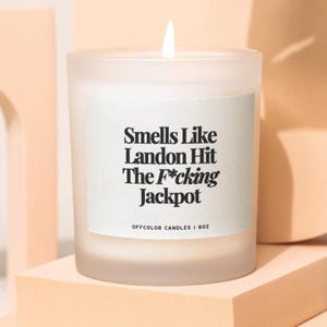 Könnte beinhalten: Eine brennende weiße Kerze in einem mattierten Glas mit einem weißen Etikett. Auf dem Etikett steht "Smells Like Landon Hit The F*cking Jackpot". Die Kerze steht auf einem pfirsichfarbenen Block. Die Kerze ist 28g.