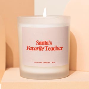 May include: A frosted glass candle with a pink label that reads "Santa's Favorite Teacher" in red text. The candle is 8 oz and has a lit white wick. The background features peach-colored geometric shapes.