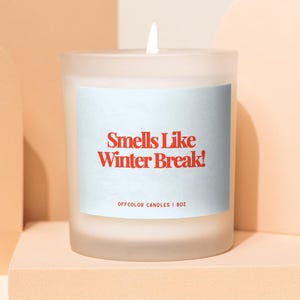 Może przedstawiać: Zapalone, matowe szklane świece z czerwonym napisem "Smells Like Winter Break!". Świeca ma 227g i stoi na platformie w kolorze brzoskwiniowym, z brzoskwiniowymi łukami w tle.