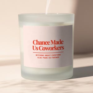 Puede incluir: Una vela encendida en un vaso esmerilado con una etiqueta rosa. La etiqueta dice "Chance Made Us Coworkers" en texto rojo, con la frase "Bitching about everyone else made us friends" debajo. Humo sube de la vela.