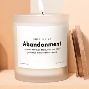 May include: A lit, frosted glass candle with the text "SMELLS LIKE Abandonment" and the phrase "notes of betrayal, tears, and how could you leave me with these people." A wooden lid rests beside the candle. Net wt. 240 g.