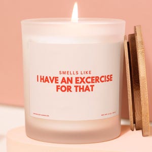 May include: A white scented candle in a frosted glass jar with a wooden lid. The candle has the text "Smells Like I Have An Excercise For That" in red. The candle is lit and sits on a pink platform.