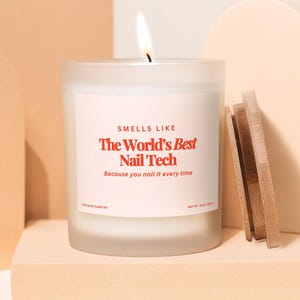 May include: A lit candle in a frosted glass jar with a wooden lid. The label reads "SMELLS LIKE The World's Best Nail Tech Because you nail it every time." The candle sits on a peach-colored surface.