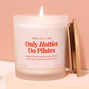 May include: A lit white candle in a frosted glass jar with a wooden lid. The label reads "Smells Like Only Hotties Do Pilates" in red text. The candle is on a light pink surface against a pink background.