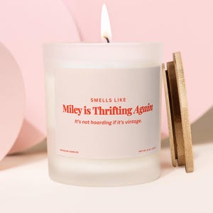Puede incluir: Una vela blanca encendida en un tarro de vidrio esmerilado con tapa de madera. La etiqueta dice "SMELLS LIKE Miley is Thrifting Again It's not hoarding if it's vintage." La vela pesa 227g.