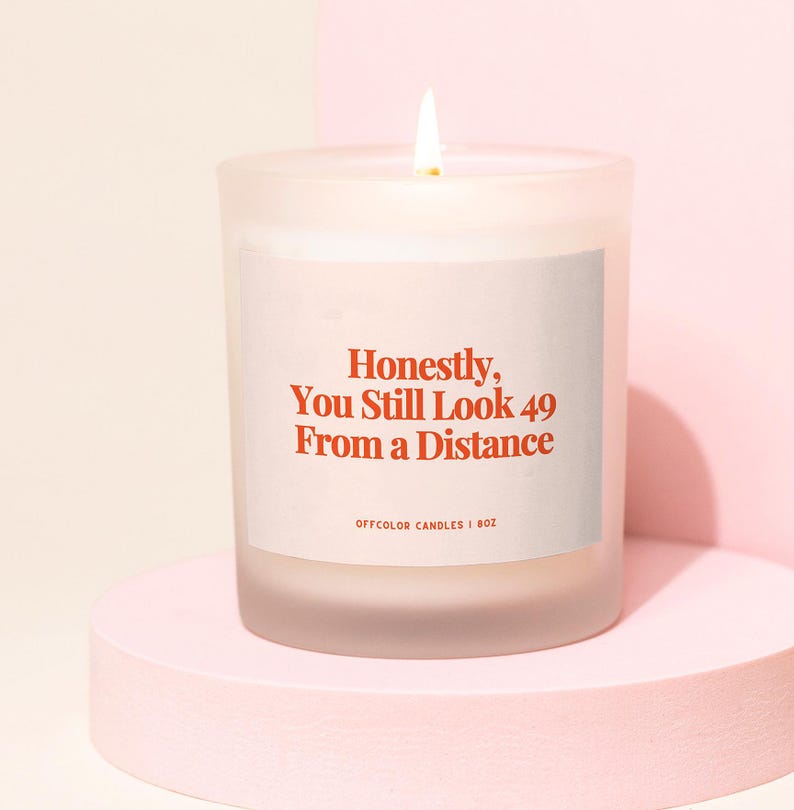 May include: A lit, frosted glass candle with the text "Honestly, You Still Look 49 From A Distance" in red. The candle is on a pink, rounded platform. The candle is 8 oz.