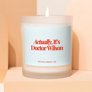 May include: A frosted glass candle with a lit wick. The candle has a light blue label with the text "Actually, It's Doctor Wilson" in red. The bottom of the label reads "OFFCOLOR CANDLES | 8OZ".