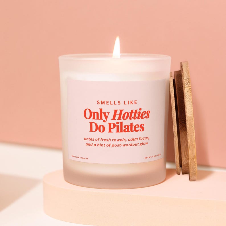 May include: A lit white candle in a frosted glass jar with a wooden lid. The label reads "Smells Like Only Hotties Do Pilates" in red text. The candle is on a light pink surface against a pink background.