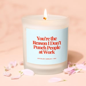 K&ouml;nnte beinhalten: Eine brennende, mattierte Glas-Kerze mit einem hellblauen Etikett. Auf dem Etikett steht "You're the Reason I Don't Punch People at Work" in roter Schrift. Die Kerze ist von rosa Bl&uuml;tenbl&auml;ttern umgeben.