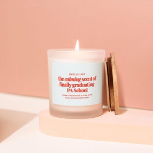 May include: A lit candle in a frosted glass jar with a wooden lid. The label reads "SMELLS LIKE the calming scent of finally graduating PA School." The candle sits on a light pink surface against a peach background.