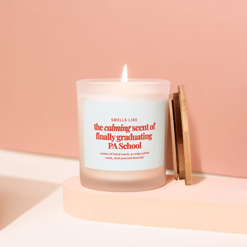 May include: A lit candle in a frosted glass jar with a wooden lid. The label reads "SMELLS LIKE the calming scent of finally graduating PA School." The candle sits on a light pink surface against a peach background.