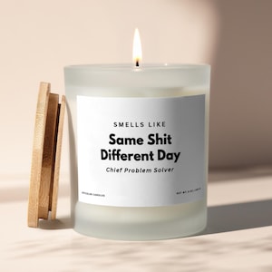 May include: A lit white candle in a frosted glass jar with a wooden lid. The candle has a white label with the text "SMELLS LIKE Same Shit Different Day Chief Problem Solver". The candle is 8 oz / 240 g.