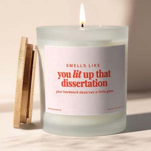 May include: A lit candle in a frosted glass jar with a wooden lid. The label reads "Smells Like you lit up that dissertation". The candle is 8 oz.