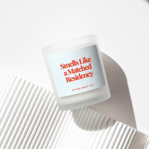 May include: White candle with a blue label that reads "Smells Like a Matched Residency" in red text. Offcolor Candles | 8oz.