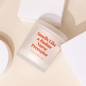 May include: A white candle with a red label that reads "Smells Like a Badass Nurse Preceptor" and "Offcolor Candles | 8oz".