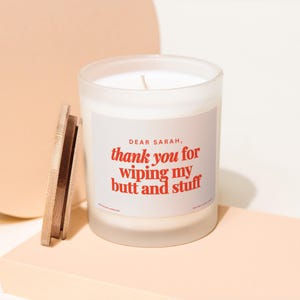May include: A white candle in a frosted glass jar with a wooden lid. The label reads "DEAR SARAH, thank you for wiping my butt and stuff" in red text. The candle sits on a light peach-colored surface.
