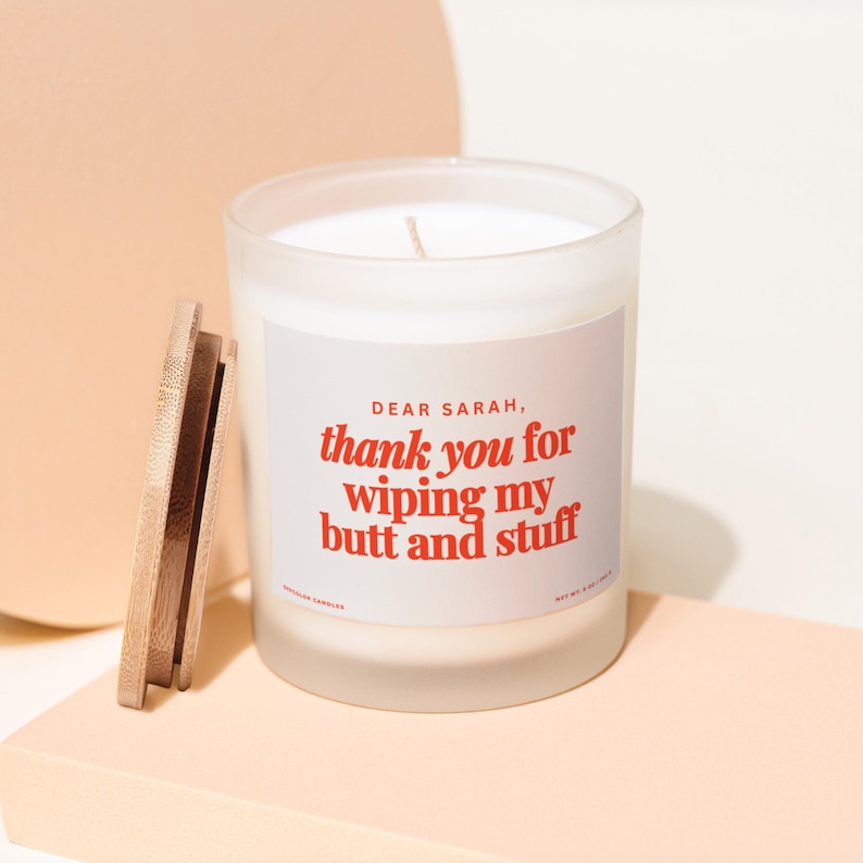 May include: A white candle in a frosted glass jar with a wooden lid. The label reads "DEAR SARAH, thank you for wiping my butt and stuff" in red text. The candle sits on a light peach-colored surface.