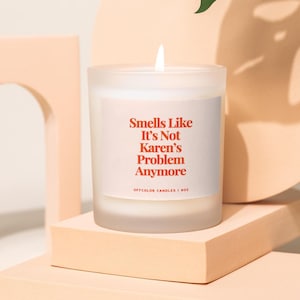 May include: A white frosted glass candle with a red label that reads "Smells Like It's Not Karen's Problem Anymore". The candle is on a light pink surface.