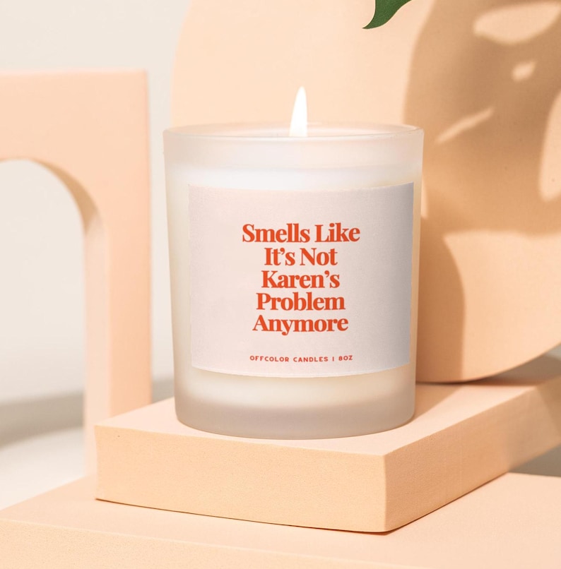 May include: A white frosted glass candle with a red label that reads "Smells Like It's Not Karen's Problem Anymore". The candle is on a light pink surface.