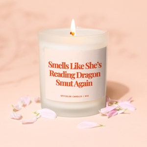 Puede incluir: Una vela de vidrio blanco esmerilado con una etiqueta que dice "Smells Like She's Reading Dragon Smut Again" y "Offcolor Candles | 8 oz". La vela está encendida y hay pétalos de flores rosas esparcidos a su alrededor.
