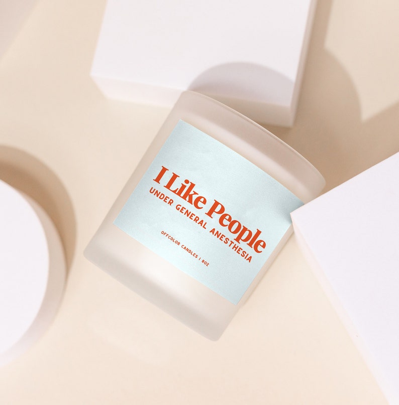 May include: A frosted glass candle with a light blue label that reads "I Like People Under General Anesthesia" in orange text. The label also says "Offcolor Candles | 8oz".