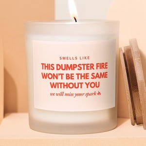 Peut inclure: Une bougie allumée dans un pot en verre dépoli avec une étiquette blanche portant le texte rouge : SMELLS LIKE THIS DUMPSTER FIRE WON'T BE THE SAME WITHOUT YOU we will miss your spark. Un couvercle en bois clair est placé à droite du pot.