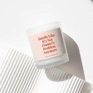May include: A white candle with a pink label that reads "Smells Like It's Not (Name)'s Problem Anymore" and "Offcolor Candles | 8oz".
