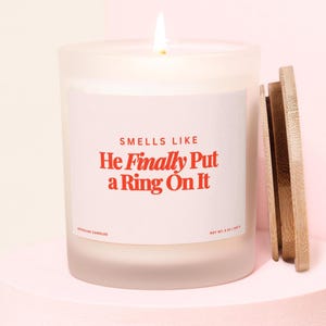 Puede incluir: Una vela blanca encendida en un tarro de vidrio esmerilado con tapa de madera. La etiqueta dice "SMELLS LIKE He Finally Put a Ring On It". La vela está sobre una superficie rosa. Peso neto 226 g.