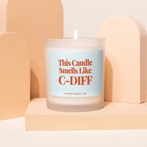 Puede incluir: Una vela de vidrio blanco esmerilado con una etiqueta azul que dice "This Candle Smells Like C-DIFF" y "OFFCOLOR CANDLES | 8OZ".