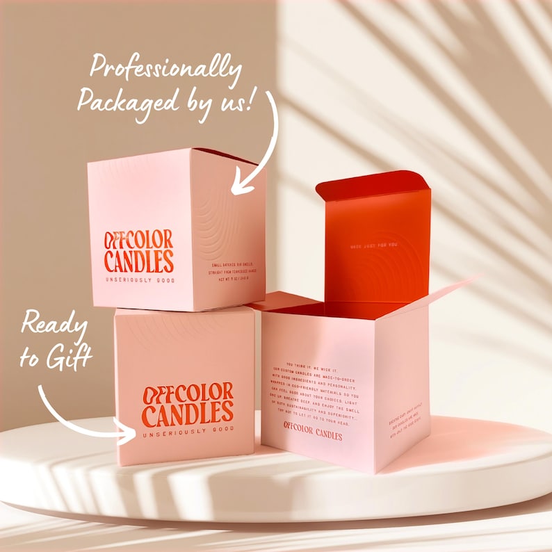 May include: Pink square boxes of Offcolor Candles, stacked on a white surface. The boxes have red text that reads "Offcolor Candles" and "Ready to Gift." One box is open, revealing a red interior. The text "Professionally Packaged by us!" is also visible.