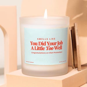 May include: A lit, frosted glass candle with a wooden lid. The candle has a light blue label with the text "Smells Like You Did Your Job A Little Too Well. Congratulations on that Promotion!" The candle sits on a peach-coloured block.