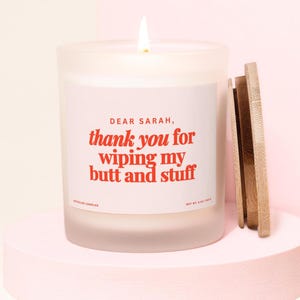 May include: A lit white candle in a frosted glass jar with a wooden lid. The label reads "DEAR SARAH, thank you for wiping my butt and stuff" in red text. The candle sits on a pink pedestal.