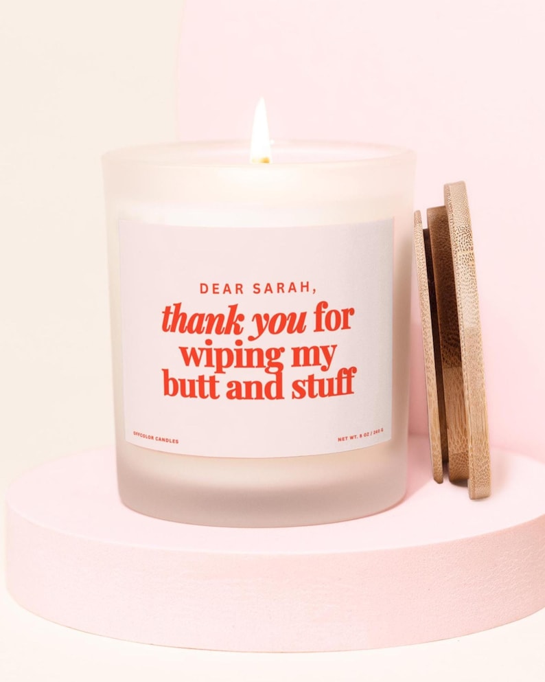 May include: A lit white candle in a frosted glass jar with a wooden lid. The label reads "DEAR SARAH, thank you for wiping my butt and stuff" in red text. The candle sits on a pink pedestal.