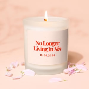 May include: A white frosted glass candle with a red label that reads "No Longer Living In Sin" and the date "10.04.2024". The candle is lit and surrounded by pink flower petals.