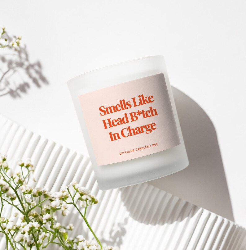 Może przedstawiać: Biała, matowa świeca w szklanym naczyniu z r&oacute;żową etykietą, na kt&oacute;rej widnieje napis "Smells Like Head B*tch In Charge" oraz "OFFCOLOR CANDLES | 8OZ".