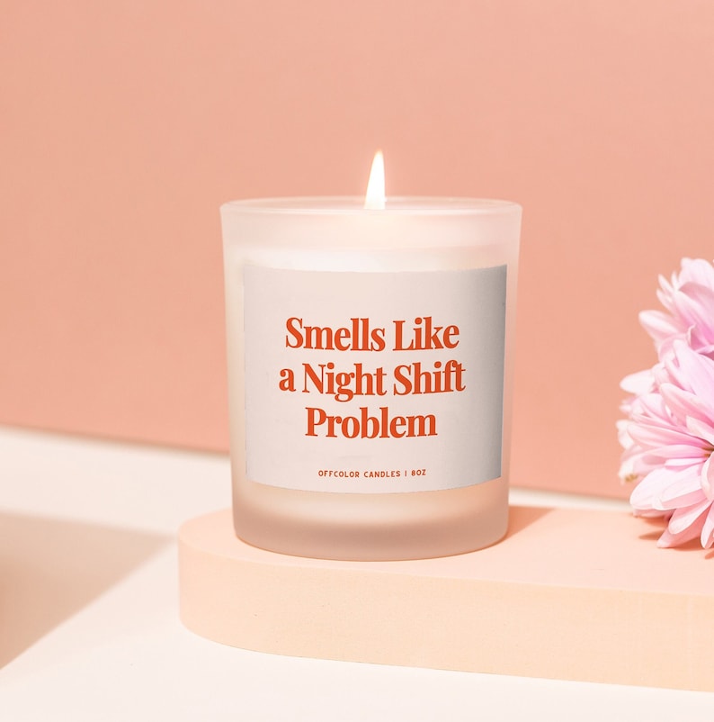 Pode incluir: Uma vela branca em um pote de vidro fosco com um r&oacute;tulo vermelho que diz "Smells Like a Night Shift Problem". A vela est&aacute; acesa e est&aacute; sobre uma superf&iacute;cie rosa claro.
