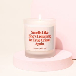 Pode incluir: Uma vela acesa de vidro fosco com o texto "Smells Like She's Listening to True Crime Again" em vermelho. A vela est&aacute; sobre um pedestal rosa circular sobre um fundo rosa claro. A etiqueta diz "OFFCOLOR CANDLES | 8OZ".