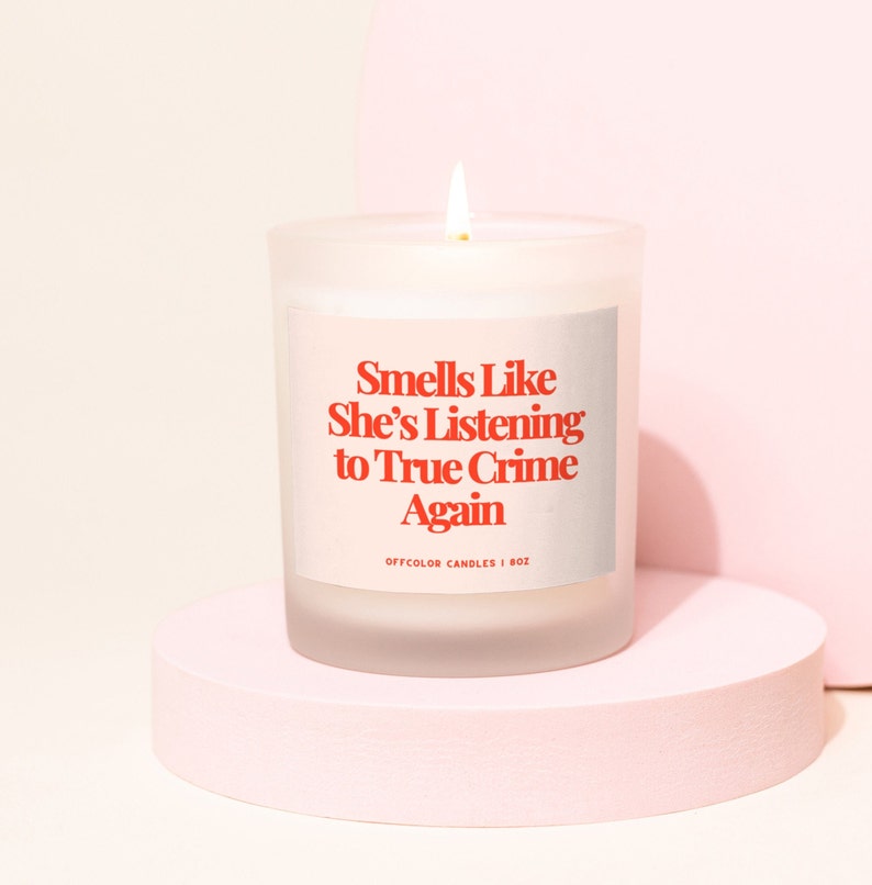 Pode incluir: Uma vela acesa de vidro fosco com o texto "Smells Like She's Listening to True Crime Again" em vermelho. A vela est&aacute; sobre um pedestal rosa circular sobre um fundo rosa claro. A etiqueta diz "OFFCOLOR CANDLES | 8OZ".