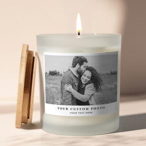 Può includere: Una candela bianca accesa in un barattolo di vetro smerigliato con coperchio in legno. La candela ha un'etichetta con una foto in bianco e nero di una coppia e il testo "YOUR CUSTOM PHOTO" e "YOUR TEXT HERE".