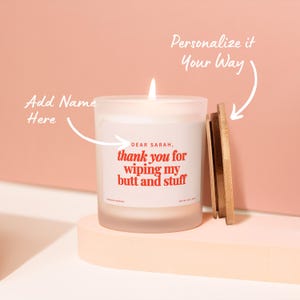 May include: A lit, frosted glass candle with a wooden lid. The candle has a label with the text "DEAR SARAH, thank you for wiping my butt and stuff" in red. The candle is on a light pink surface.
