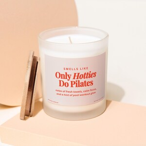 May include: A frosted glass candle with a wooden lid. The candle has a label that reads "SMELLS LIKE Only Hotties Do Pilates" with scent notes of fresh towels, calm focus, and a hint of post-workout glow.