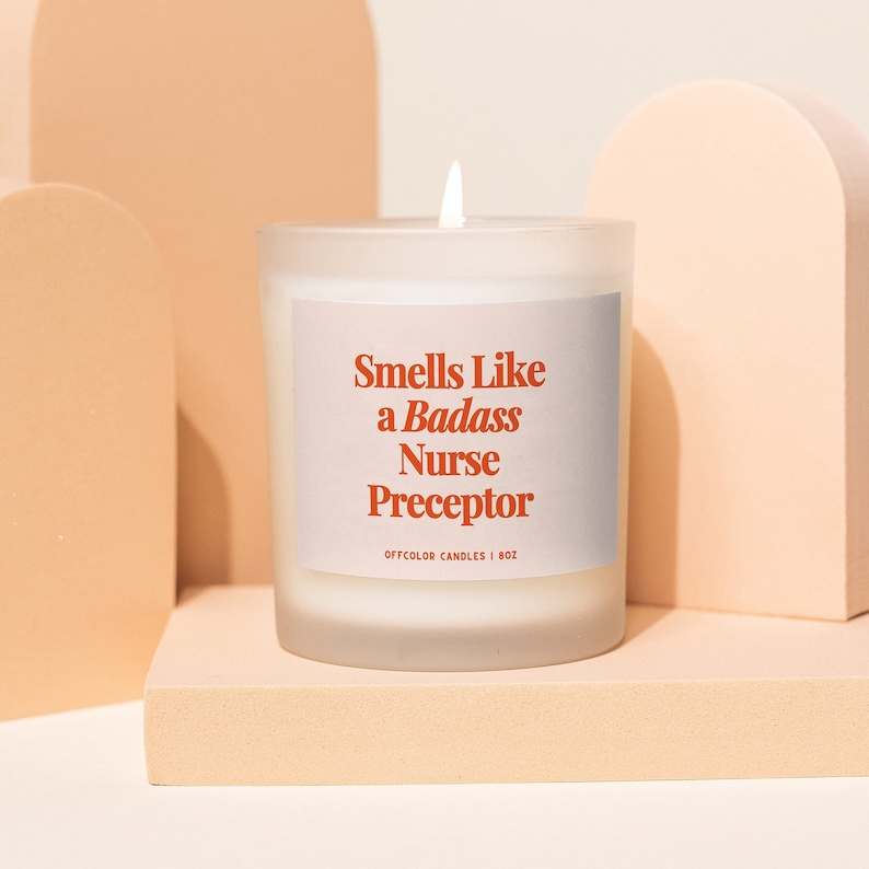 May include: White frosted glass candle with a red label that reads "Smells Like a Badass Nurse Preceptor". The label also includes the text "Offcolor Candles | 8 oz".