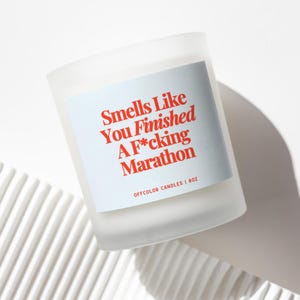 May include: A frosted glass candle with a light blue label featuring the text "Smells Like You Finished A F*cking Marathon" in red. The candle is 8 oz and from Offcolor Candles. The candle sits on a white, textured surface.