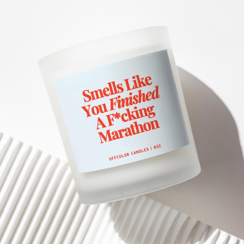 May include: A frosted glass candle with a light blue label featuring the text "Smells Like You Finished A F*cking Marathon" in red. The candle is 8 oz and from Offcolor Candles. The candle sits on a white, textured surface.