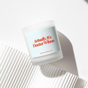 May include: A frosted glass candle with a light blue label. The label reads "Actually, It's Doctor Wilson" in red text. The candle is 1 oz. and sits on a white, textured surface. The candle is a home decor item.