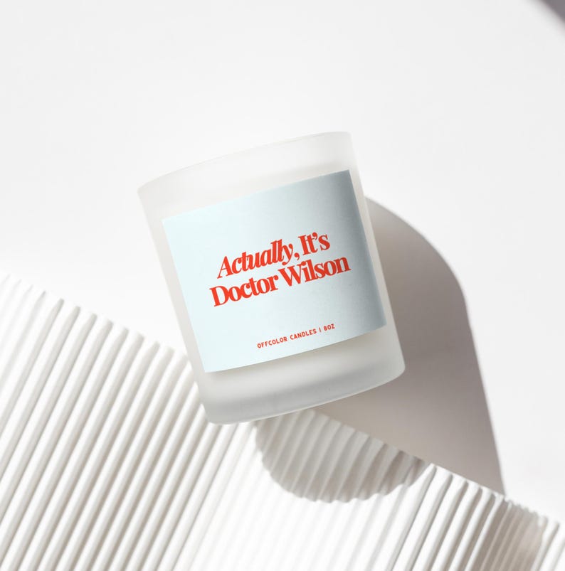 May include: A frosted glass candle with a light blue label. The label reads "Actually, It's Doctor Wilson" in red text. The candle is 1 oz. and sits on a white, textured surface. The candle is a home decor item.