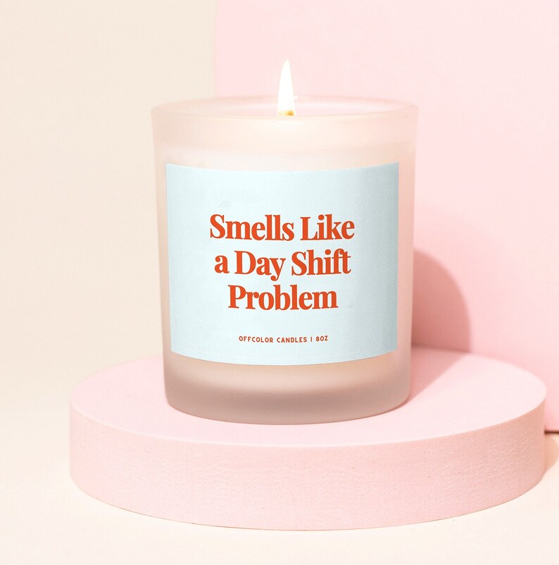 May include: A white frosted glass candle with a blue label that reads "Smells Like a Day Shift Problem" and "Offcolor Candles | 8oz".
