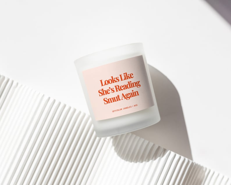May include: A white frosted glass candle with a pink label that reads "Looks Like She's Reading Smut Again" and "Offcolor Candles | 8oz".